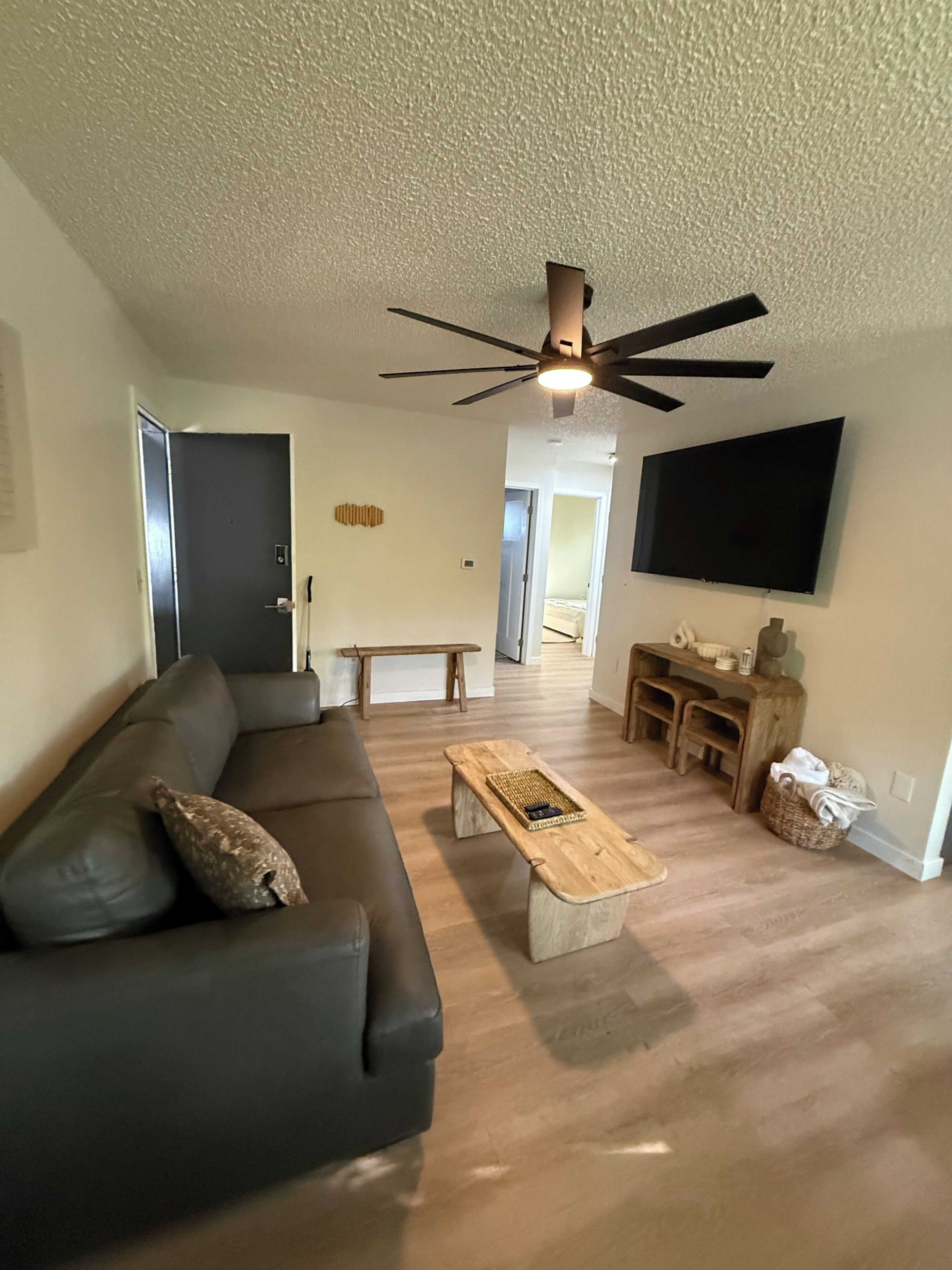 Una sala de estar con un sofá de color oscuro, una mesa de centro de madera, un ventilador de techo y un televisor de pantalla plana montado en la pared.