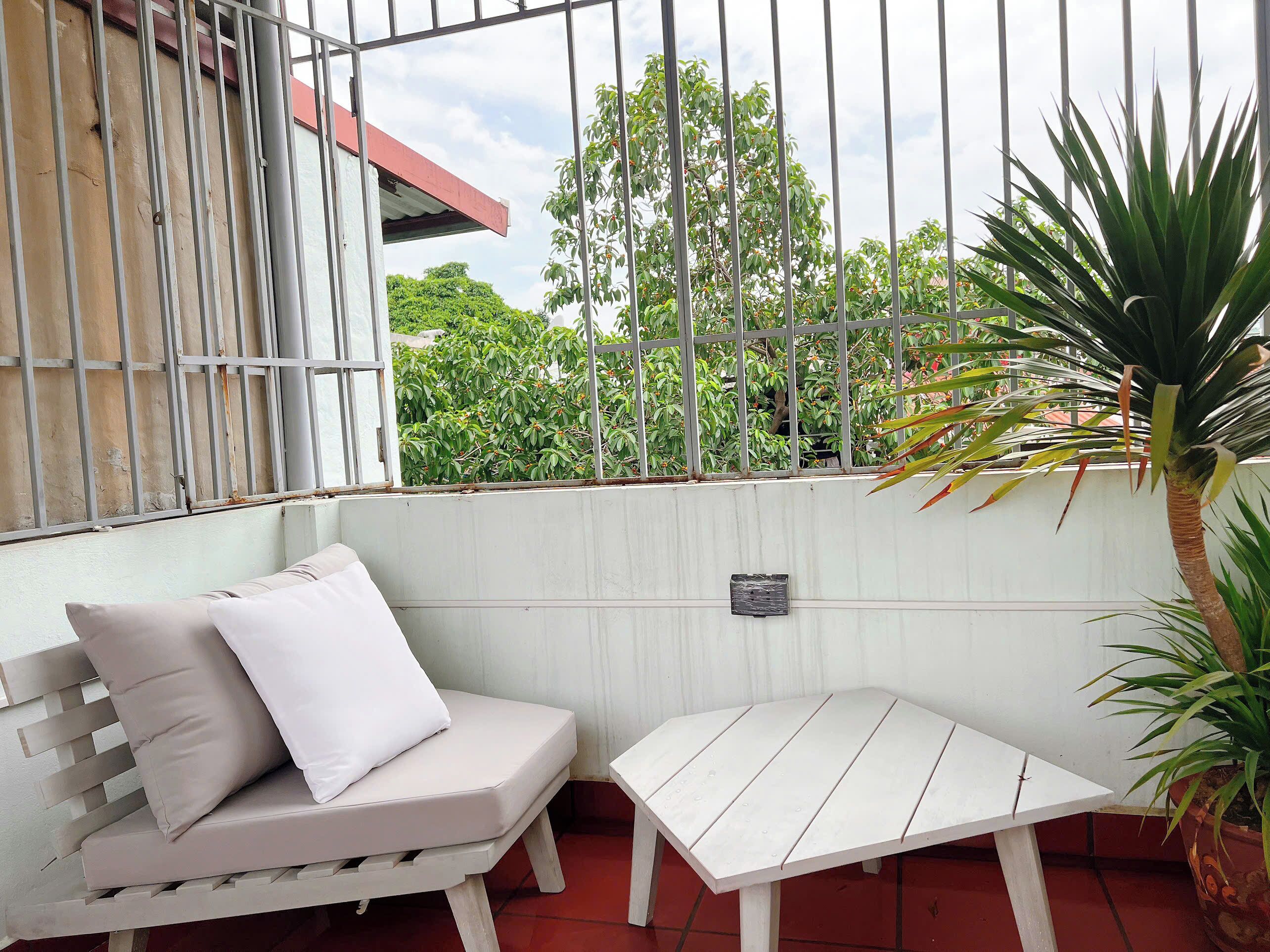 A white outdoor seating area with a bench and a small table, surrounded by a metal fence and greenery.