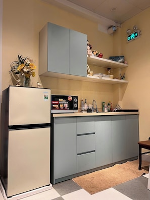 Cocina compacta con refrigerador, microondas y pileta. Hay armarios sobre la encimera y un pequeño estante con diversos objetos.
