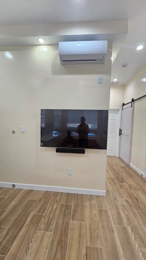 Una habitación con un televisor de pantalla plana grande montado en la pared, luces empotradas en el techo, piso de madera y una puerta blanca.