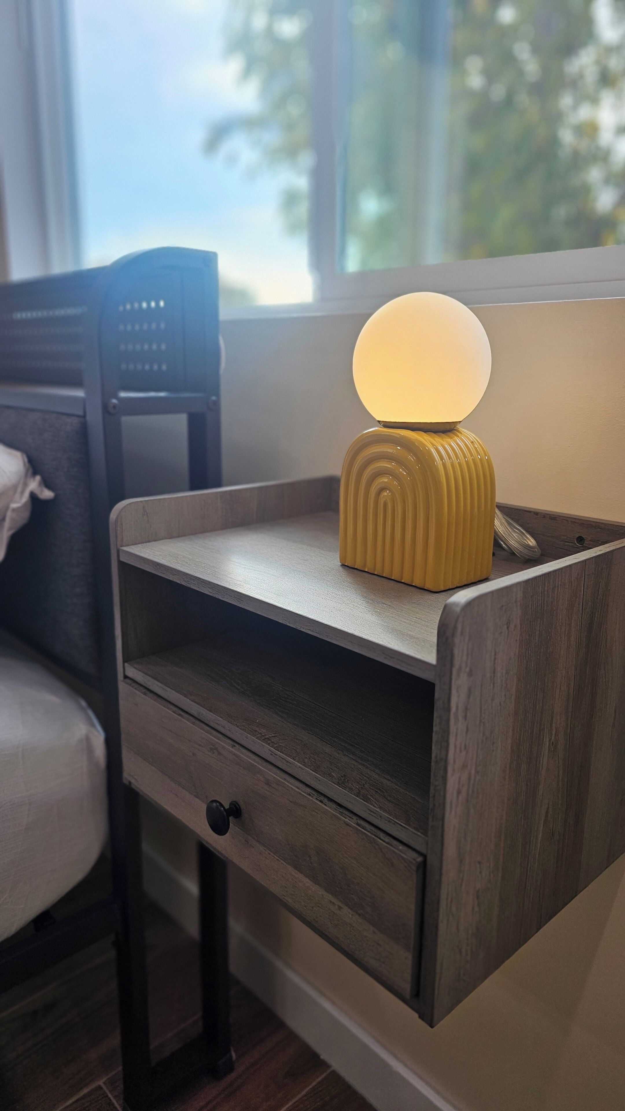 A bedside table with a lamp, a drawer, and a chair in the background.