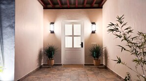 Un pasillo con una puerta blanca, dos luces empotradas en la pared y plantas en macetas a ambos lados.