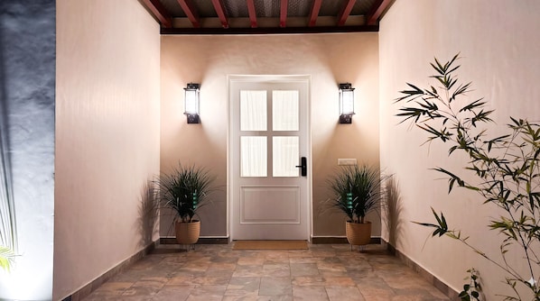 A hallway with a white door, two wall-mounted lights, and potted plants on either side.