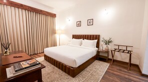 A hotel room with a large bed, bedside tables, a nightstand, a lamp, a wooden bench, and a window with curtains.