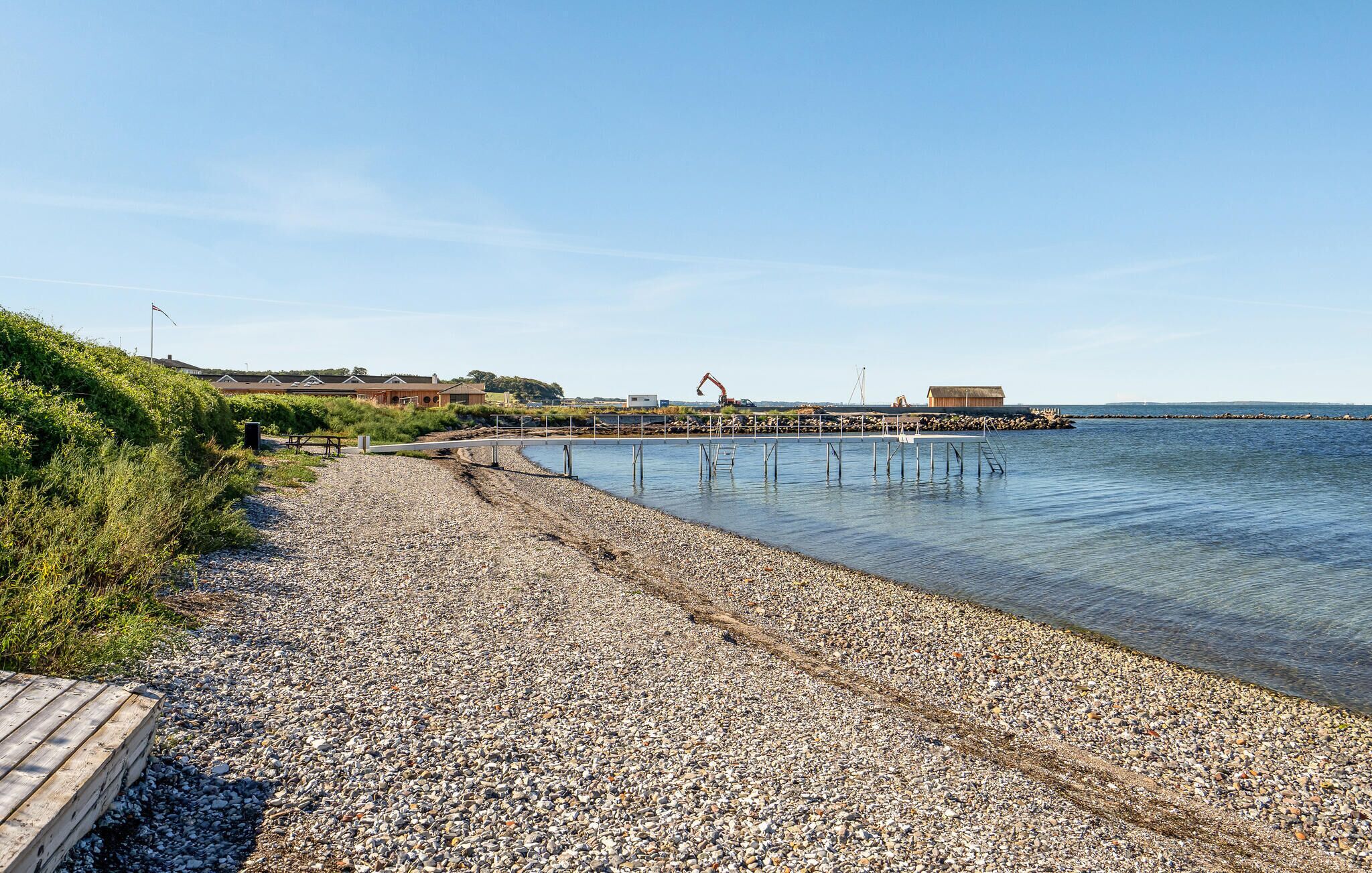 Une plage de galets avec une promenade en bois qui mène à une jetée s’avançant dans des eaux calmes.