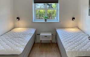 Et lille, godt oplyst værelse med to senge, et vindue med udsigt til grønne områder, og et lille hvidt natbord.
