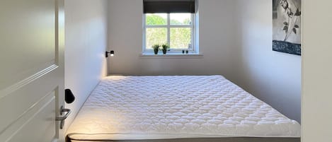 A bedroom with a bed, a window with plants, and a wall-mounted picture.