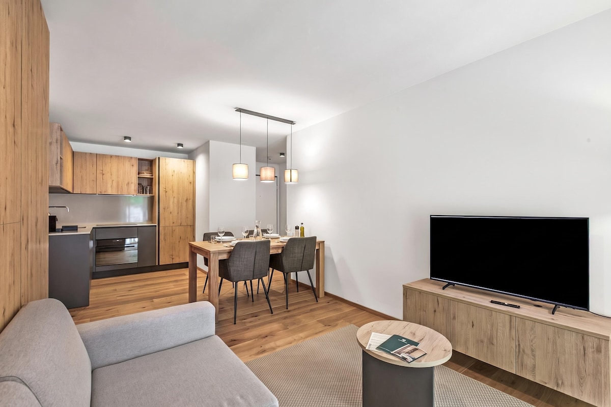 A modern living room with a flat-screen TV, a grey sofa, and a dining area with a wooden table and chairs.