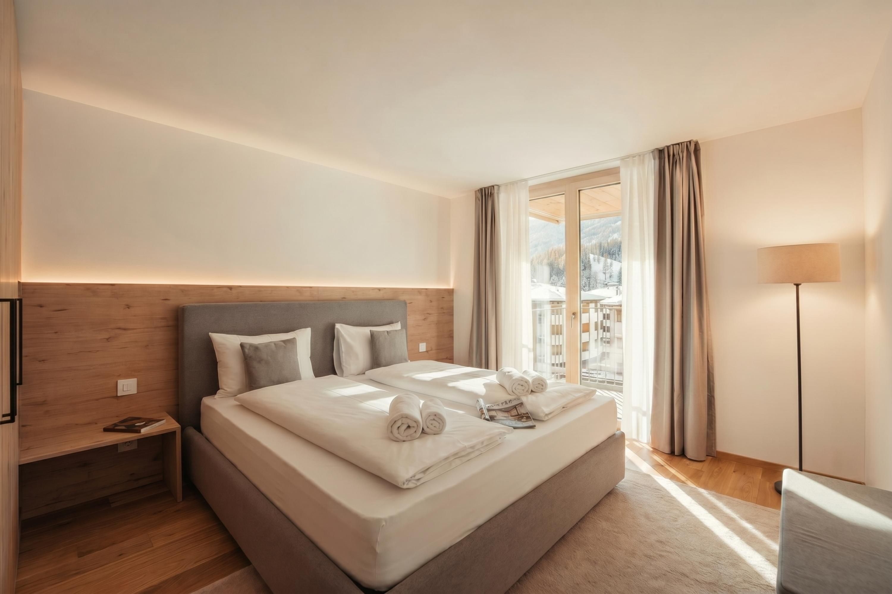 Ein Hotelzimmer mit einem großen Bett, einem Nachttisch, einer Lampe und Blick auf eine verschneite Landschaft durch ein Fenster.