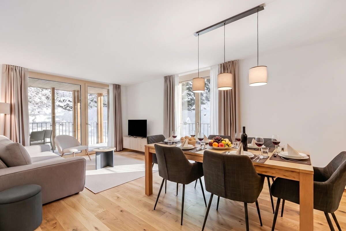 A modern living room with a dining area, large windows, and a flat-screen TV.