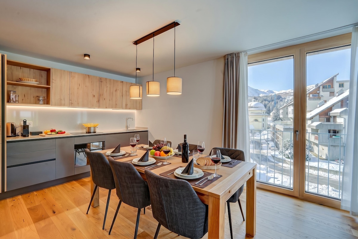 A modern kitchen with wooden cabinets, a dining area with a table set for two, and a large window offering a view of a snowy landscape.
