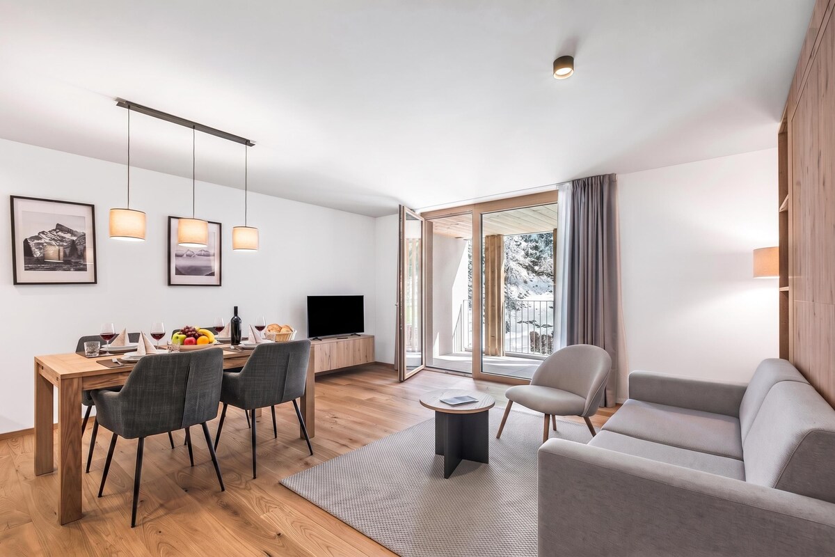 A modern living room with a dining area, a flat-screen TV, and a large window offering a view of the outdoors.