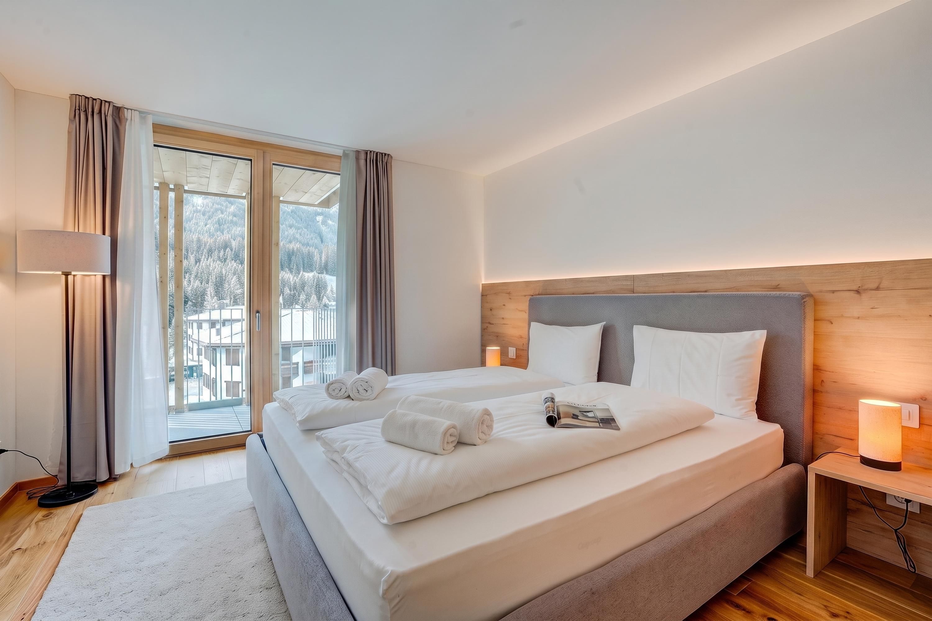 Ein Hotelzimmer mit einem grossen Bett, einem Holzkopfstück, einem Nachttisch mit Lampe und Blick auf eine verschneite Landschaft durch das Fenster.