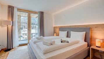 Ein Hotelzimmer mit einem grossen Bett, einem Holzkopfstück, einem Nachttisch mit Lampe und Blick auf eine verschneite Landschaft durch das Fenster.
