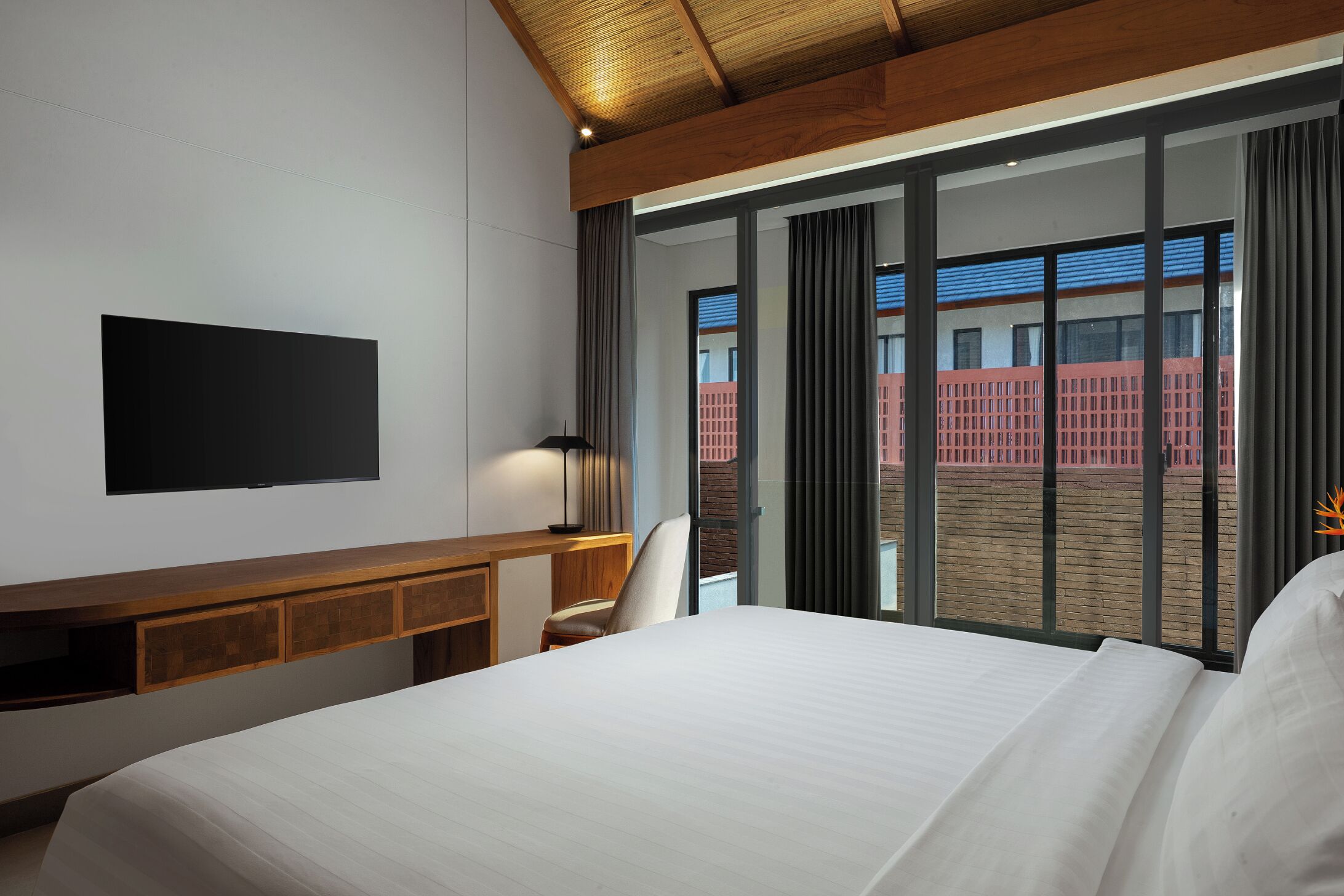 Habitación de hotel con cama, escritorio, silla, televisor y vistas a un edificio a través de amplios ventanales.