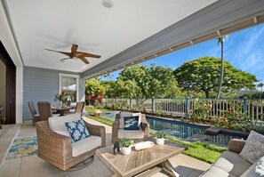 A covered outdoor seating area with wicker furniture, a pool, and a view of a lush garden. - Island Time-Luxury Golf Front Home with Pool/Spa (Kailua-Kona)