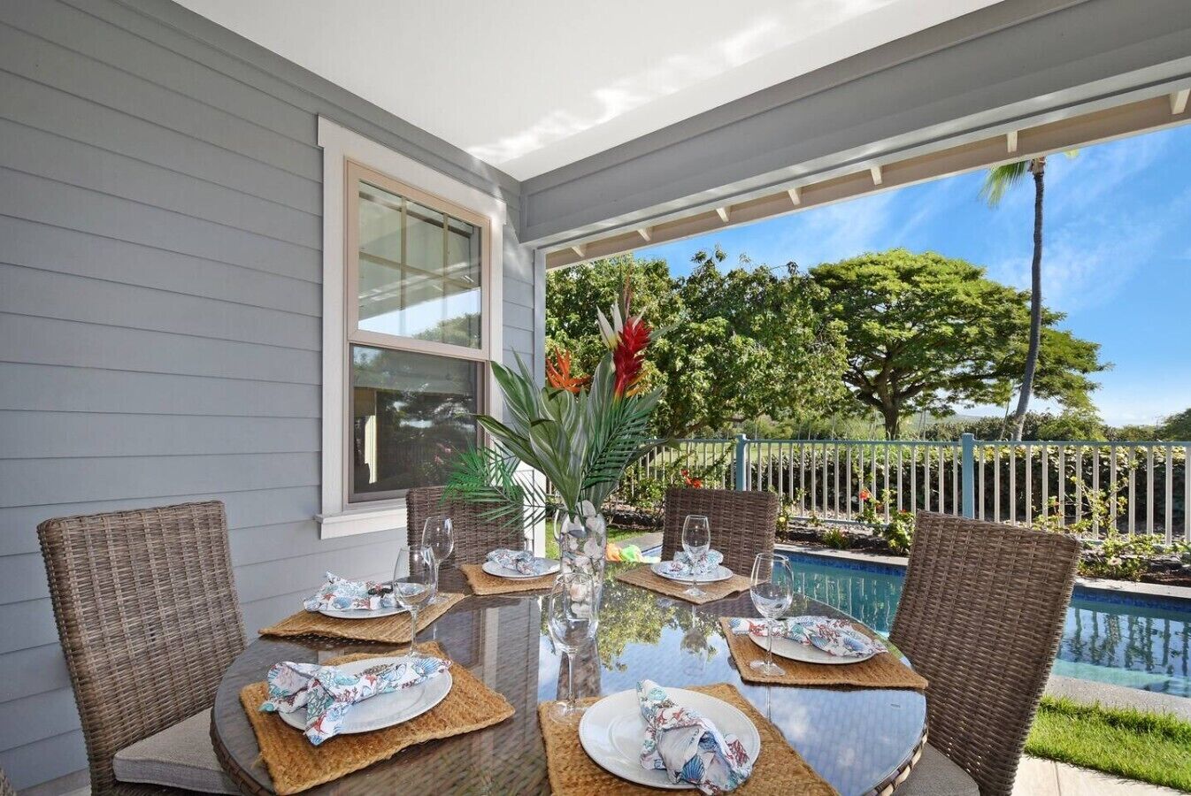 An outdoor dining area with a glass table, wicker chairs, and a view of a swimming pool and trees.