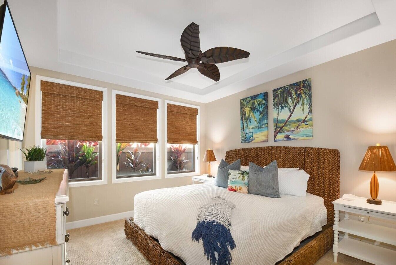 A bedroom with a bed, a ceiling fan, a TV, and a window with a view of plants.