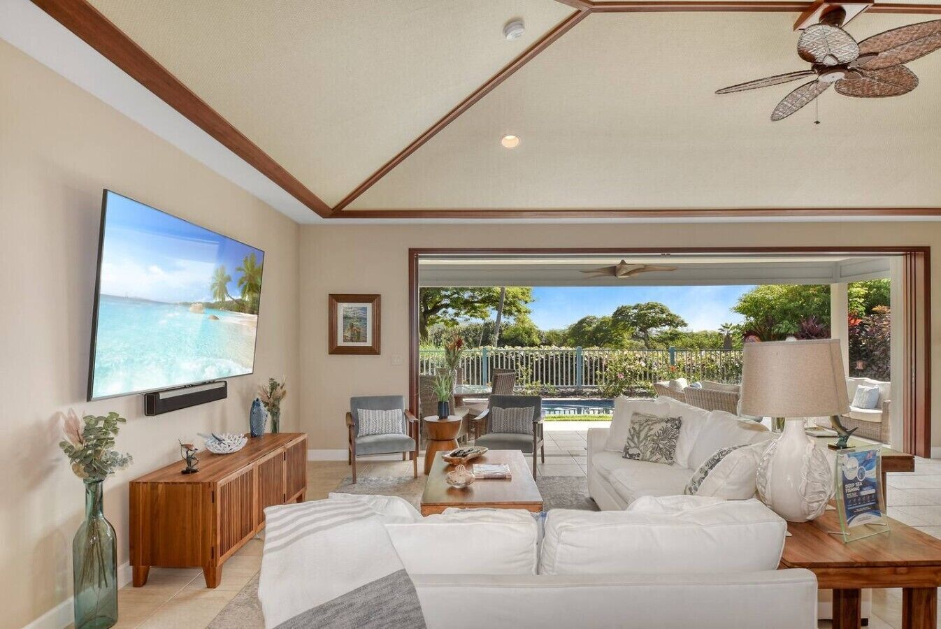 Un salon spacieux doté d’un grand téléviseur à écran plat, d’un ventilateur de plafond et offrant une vue sur une piscine et un jardin, grâce à une porte vitrée.