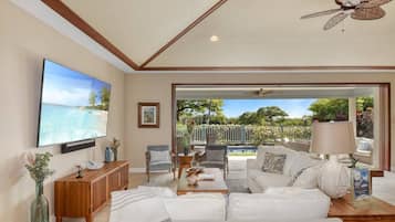A spacious living room with a large flat-screen TV, a ceiling fan, and a view of a pool and garden through a glass door.