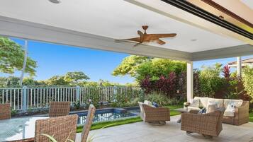 A covered outdoor area with wicker furniture, a ceiling fan, and a view of a pool and garden.