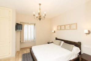 A bedroom with a bed, bedside tables, a TV mounted on the wall, a chandelier, and a window with curtains.