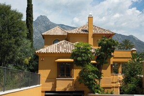 Un edificio amarillo con tejado de baldosas, rodeado de vegetación y montañas al fondo.