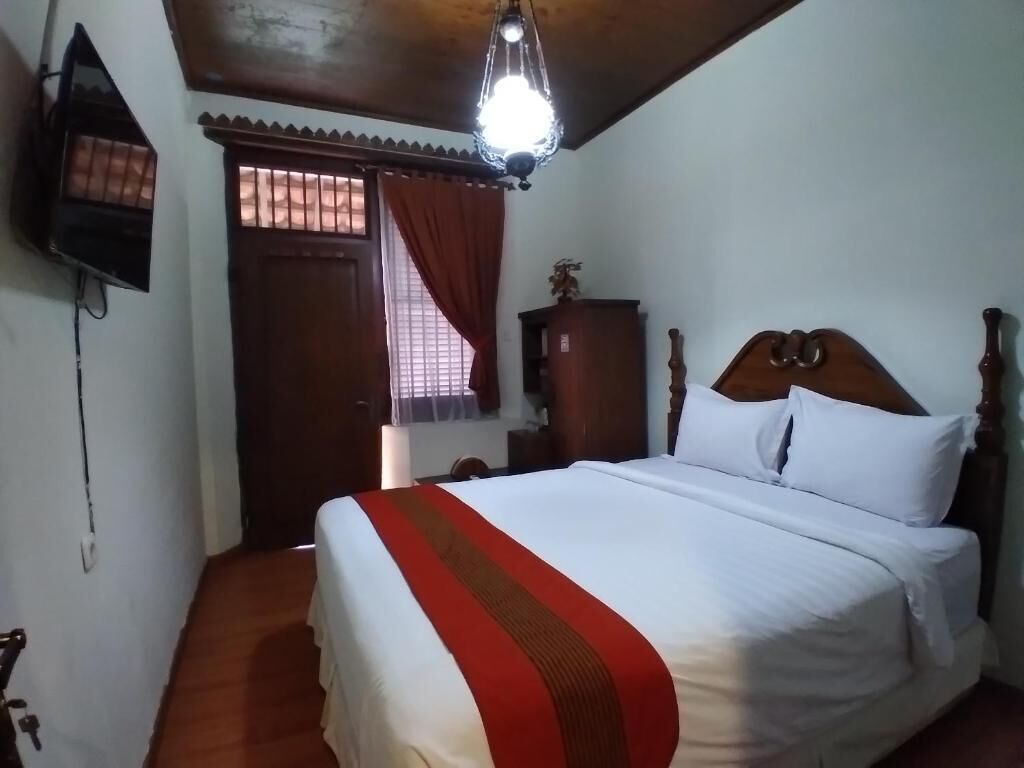 Una habitación de hotel con una cama de madera, una mesita de noche, un televisor montado en la pared y una ventana con cortinas.