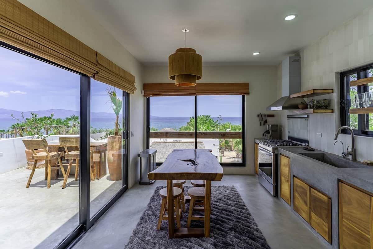 A modern kitchen with a large island, wooden chairs, and a view of the ocean.