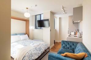 A bedroom with a bed, a television, a blue sofa, and a kitchen area with a kettle. - Studio for 2 in Central Te Aro Stay! (Wellington)
