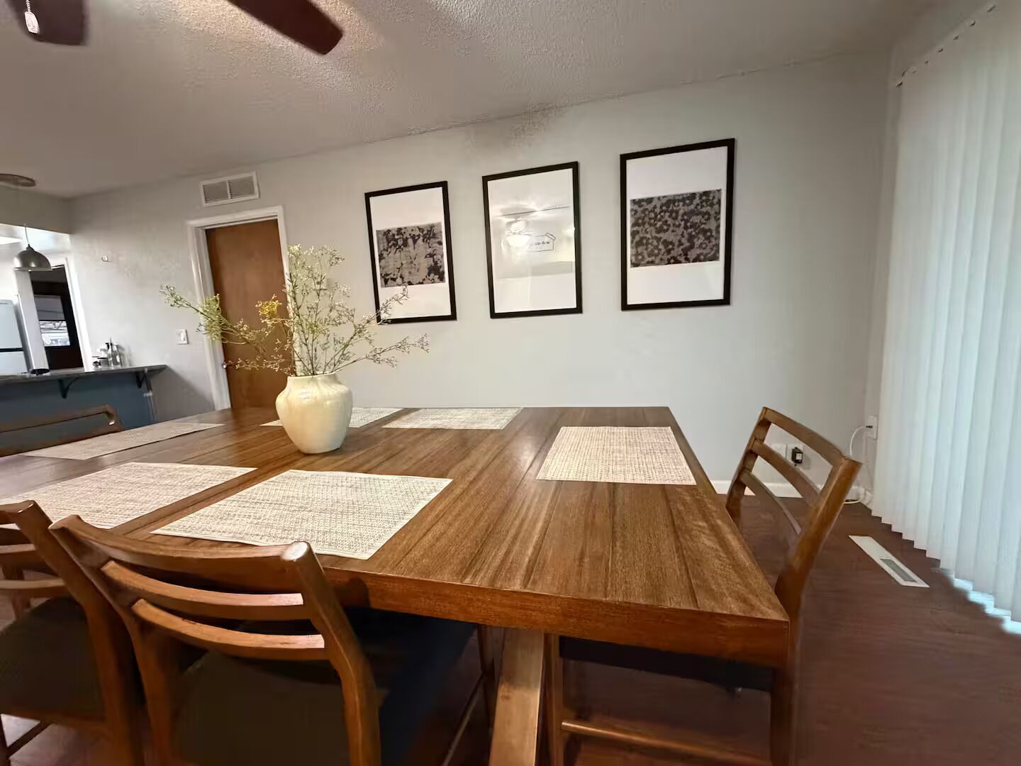 A dining area with a wooden table, chairs, and a vase with flowers. There are three framed pictures on the wall.