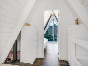 Ein schmaler Korridor mit weißen Wänden und Holzboden, der in ein Schlafzimmer mit Bett und Fenster führt.