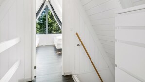 Una habitación estrecha en el ático, con paneles blancos, techo inclinado, una lámpara colgante y vistas al exterior con vegetación.