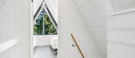 A narrow, white-paneled attic room with a sloped ceiling, a single hanging light fixture, and a view of greenery outside.