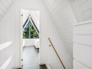 Ein schmales Dachzimmer mit weißen Wandverkleidungen, schrägen Wänden, einer einzelnen Pendelleuchte und Blick auf Grünfläche draußen.