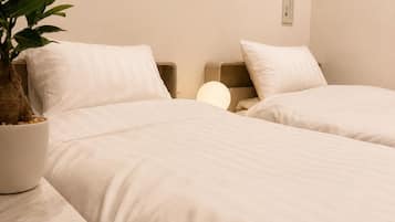 A hotel room with two beds, a bedside table with a plant, a wall-mounted lamp, and a window with curtains.