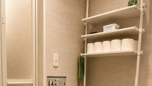Un baño con inodoro, estante con papel higiénico y escoba.
