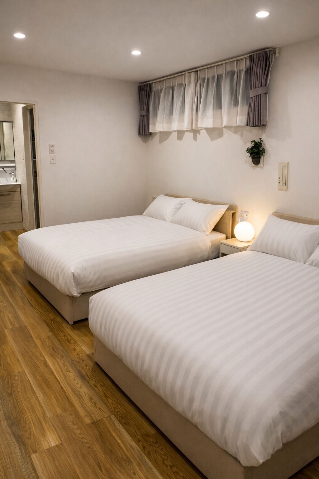 Quarto de hotel com duas camas, uma mesa de cabeceira com abajur, janela com cortinas e piso de madeira.