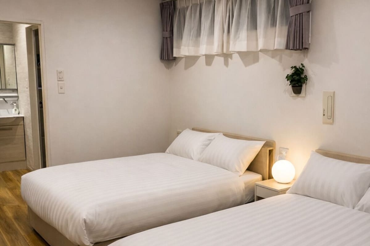 Habitación de hotel con dos camas, una mesita de noche con lámpara, una ventana con cortinas y piso de madera.