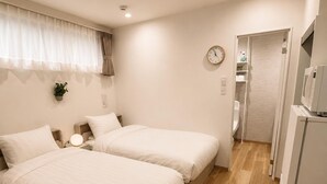 Habitación de hotel con dos camas, una mesita de noche con lámpara, un reloj en la pared y un refrigerador.