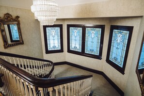 A staircase with a dark wooden handrail and white balusters, leading to a landing with a chandelier and stained glass windows.