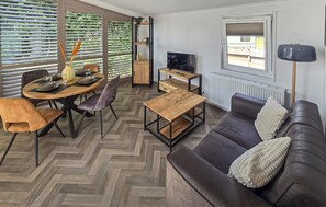 Un salon moderne avec un parquet en chevrons, une table à manger en bois avec des chaises, un canapé avec des coussins, un téléviseur et une fenêtre donnant sur l’extérieur.
