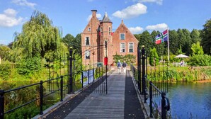 A wooden bridge leading to a historic brick building with a flag, surrounded by greenery and a calm water body. - Gorgeous home in Oude Pekela with sauna (Oude Pekela)