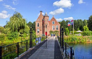 A wooden bridge leading to a historic brick building with a flag, surrounded by greenery and a calm water body. - Gorgeous home in Oude Pekela with sauna (Oude Pekela)