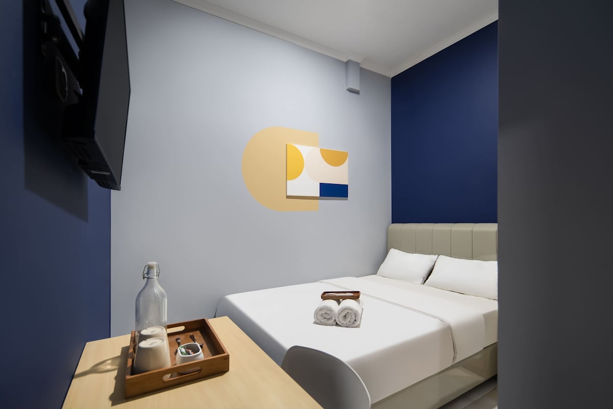 A modern hotel room with a bed, a wooden side table with toiletries, a wall-mounted TV, and a unique geometric wall art piece.