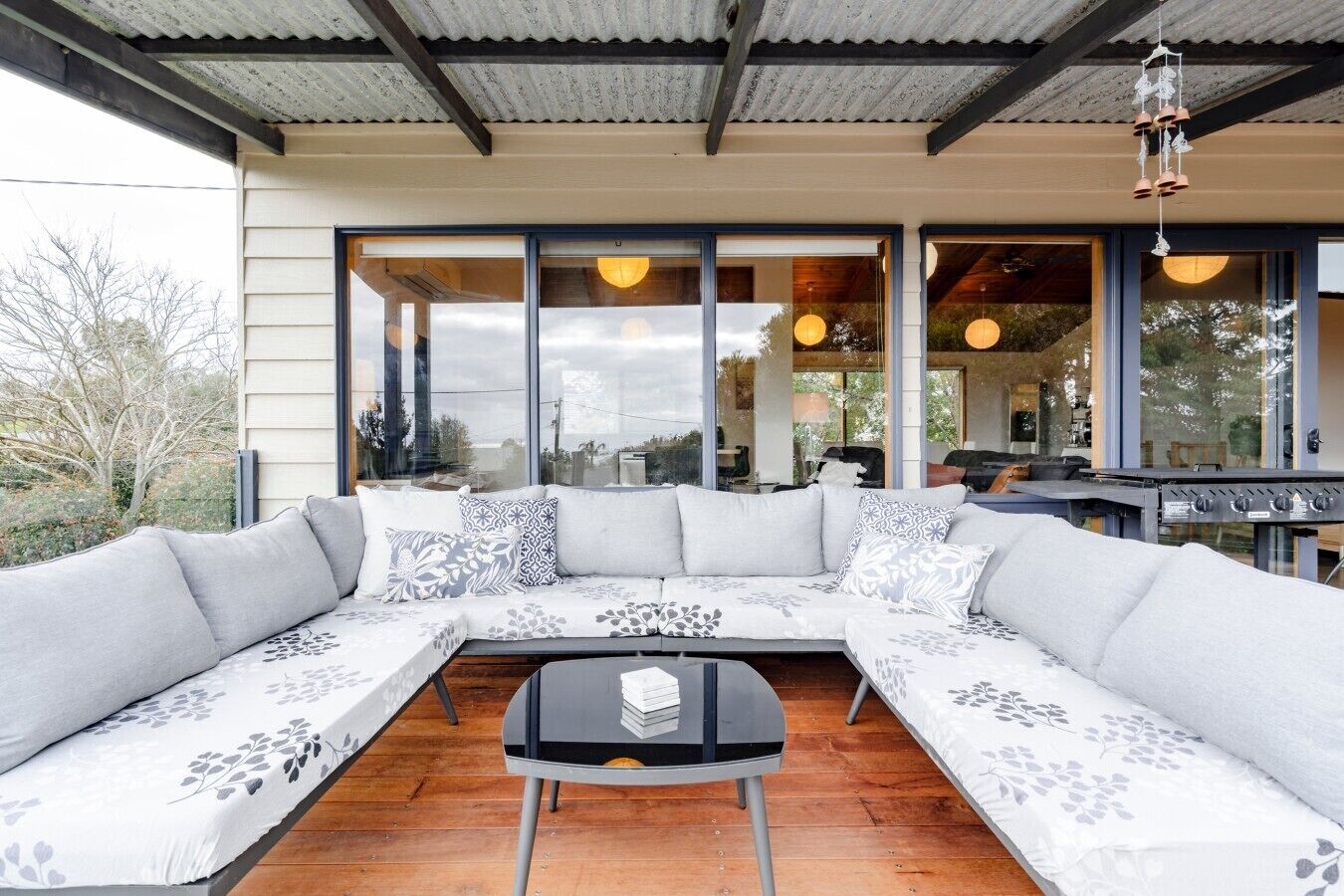 A modern outdoor seating area with a sectional sofa and a small table, large windows, and a metal roof structure.
