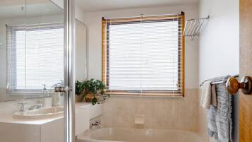 A modern bathroom with a glass shower enclosure, a bathtub, a vanity with a sink, and a window with blinds.