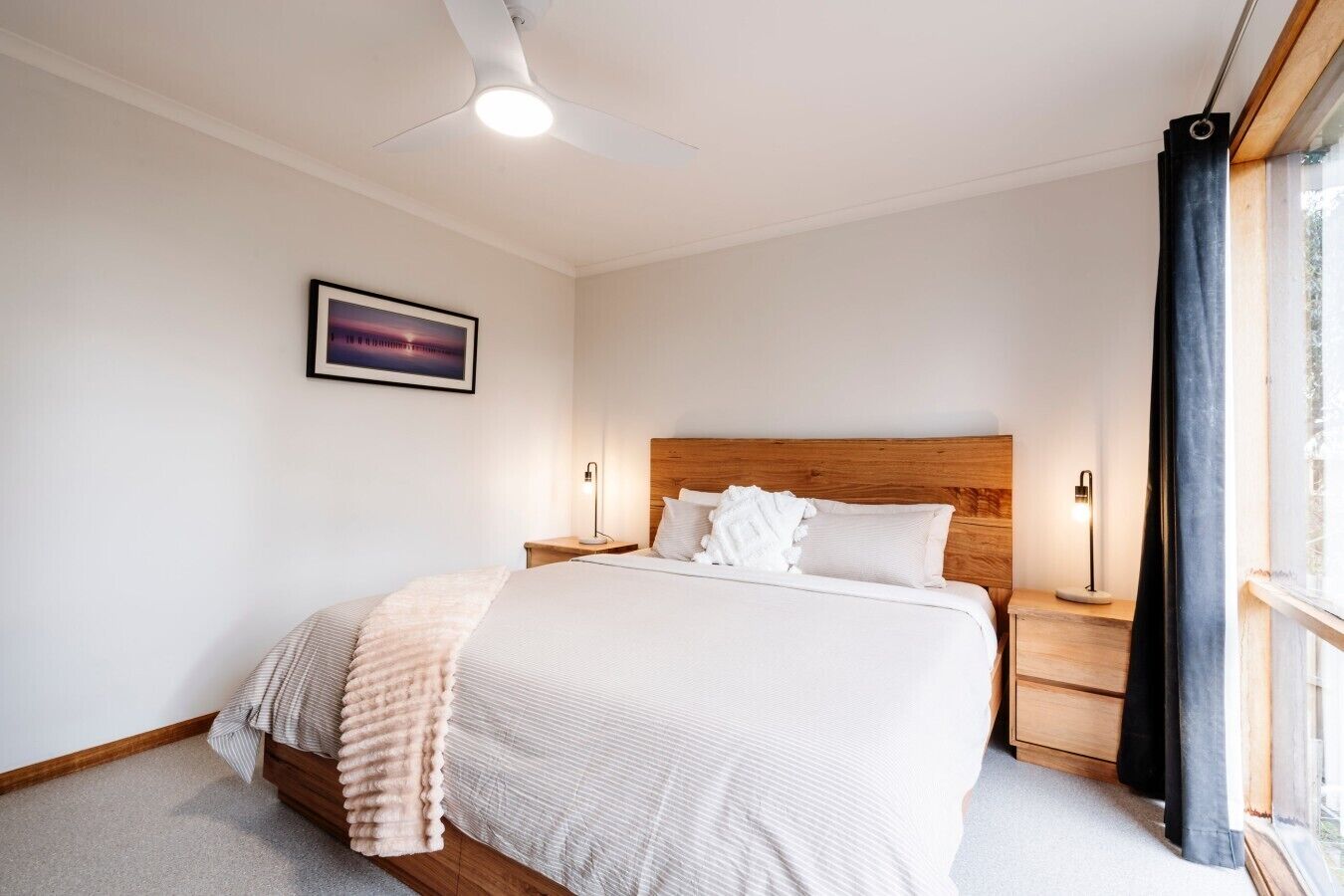 A bedroom with a bed, bedside tables, a ceiling fan, a window with curtains, and a framed picture on the wall.