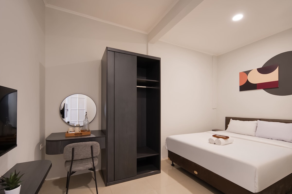 A modern hotel room with a bed, a desk with a chair, a wardrobe, a mirror, and a wall art piece.