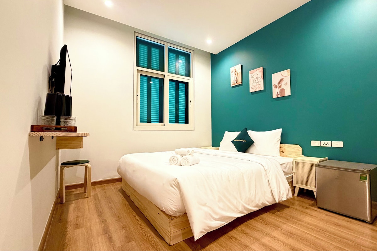 Quarto de hotel com uma cama, uma pequena escrivaninha, televisão e janela com persianas.
